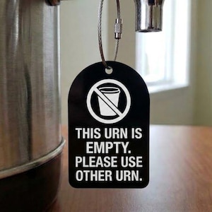 May include: A black rectangular tag with rounded top corners hangs from a silver metal urn by a wire loop. The tag features a white graphic of a cup with a diagonal line through it and the text: THIS URN IS EMPTY. PLEASE USE OTHER URN.
