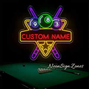 May include: Neon sign with a billiards theme. The sign features a star and cue sticks, with the text "CUSTOM NAME". The sign has a neon glow in yellow, red, green, and purple. A pool table is in the foreground.