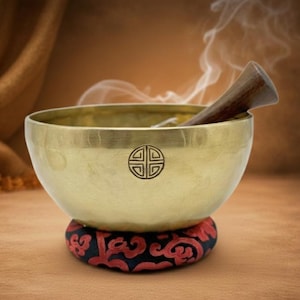 May include: A brass singing bowl with a wooden striker, set on a decorative red and black cushion. The bowl features an etched symbol and emits a wisp of smoke. The background is a warm, neutral tone.