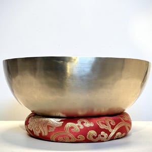 May include: A smooth, metallic, gold-toned singing bowl with a wide, open rim. The bowl rests securely on a circular, padded cushion featuring a vibrant red fabric base decorated with an intricate, swirling gold-colored floral pattern.