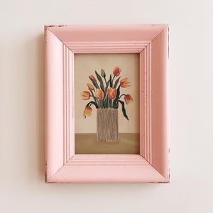 Könnte beinhalten: Ein rosa bemalter Holzrahmen mit einer einfachen Illustration eines Straußes orangefarbener Tulpen in einer weißen Vase. Das Kunstwerk befindet sich auf einem cremefarbenen Hintergrund.