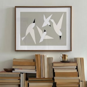 May include: A framed wall art print featuring three stylized white birds with black beaks against a solid light gray background. The artwork is displayed in a thin brown wooden frame, positioned above a large, disorganized collection of stacked books.