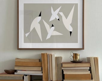 May include: A framed wall art print featuring three stylised white birds with black beaks against a solid light grey background. The artwork is displayed in a thin brown wooden frame, positioned above a large, disorganised collection of stacked books.