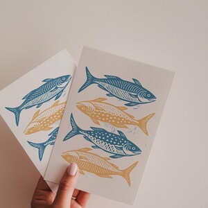 K&ouml;nnte beinhalten: Zwei Karten mit einem blauen und gelben Fischdruck. Die Fische sind stilisiert und haben ein klotziges Design. Die Karten werden in einer Hand gehalten.