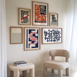 May include: A living room interior with a white wall featuring a collection of framed posters with black and red text. The posters are in German. A beige armchair with a fluffy texture sits in front of the wall. A small beige table with a book on top sits in front of the chair.
