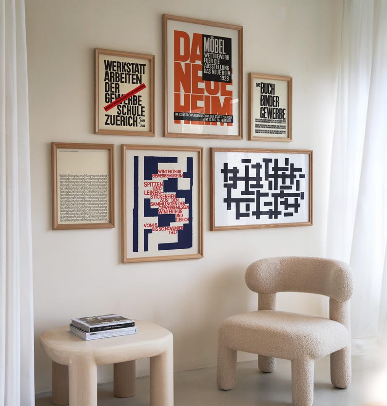 May include: A living room interior with a white wall featuring a collection of framed posters with black and red text. The posters are in German. A beige armchair with a fluffy texture sits in front of the wall. A small beige table with a book on top sits in front of the chair.