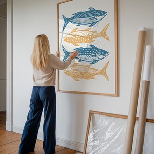 Op de afbeelding: Een vrouw hangt een ingelijst drukwerk van vier gestileerde vissen aan een witte muur. De vissen zijn in afwisselende kleuren blauw en geel. Het drukwerk is in een houten lijst.