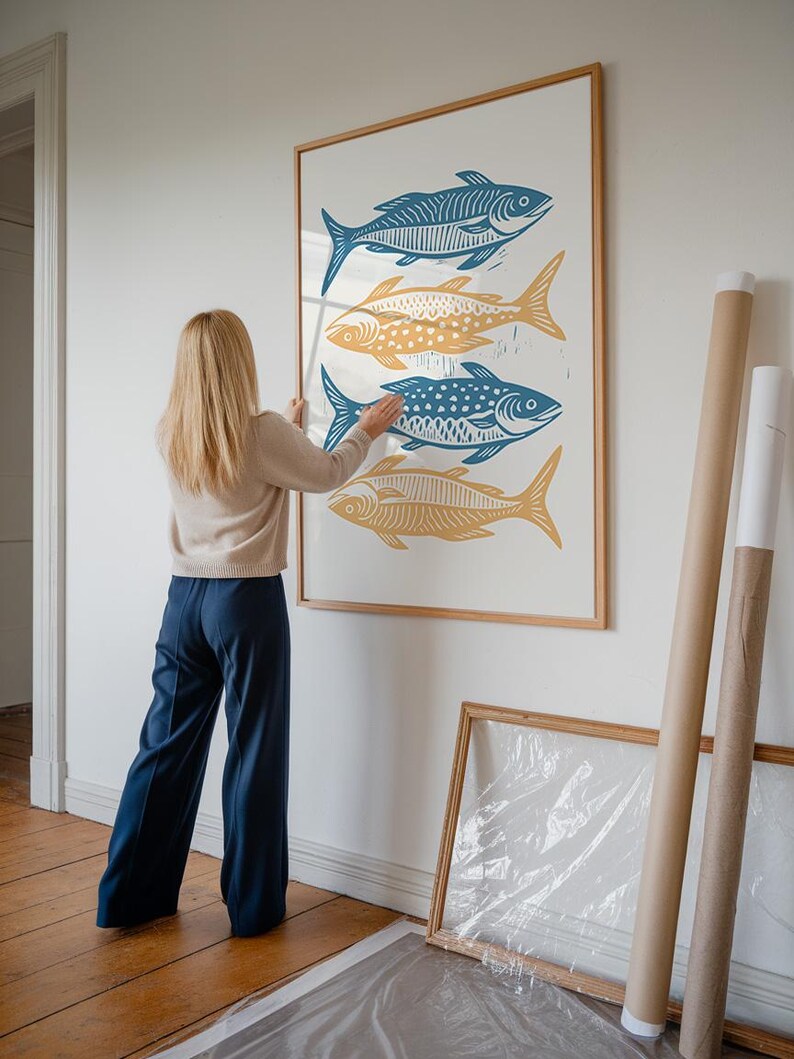Op de afbeelding: Een vrouw hangt een ingelijst drukwerk van vier gestileerde vissen aan een witte muur. De vissen zijn in afwisselende kleuren blauw en geel. Het drukwerk is in een houten lijst.