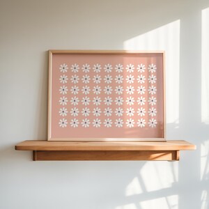 May include: A framed print with a pink background and a pattern of white daisies. The print is hanging on a wooden shelf.