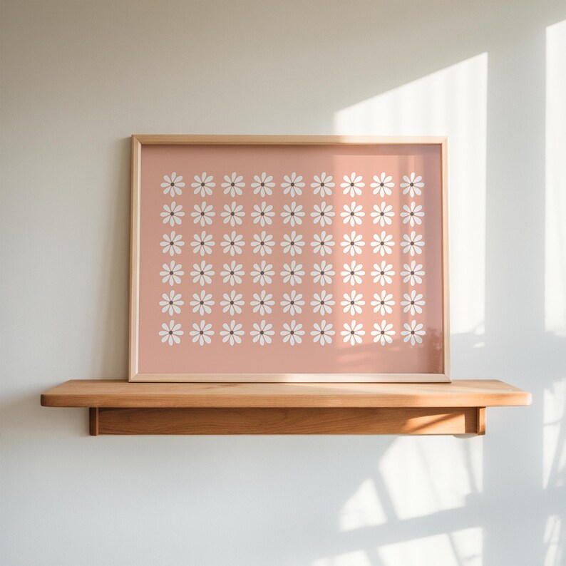 May include: A framed print with a pink background and a pattern of white daisies. The print is hanging on a wooden shelf.