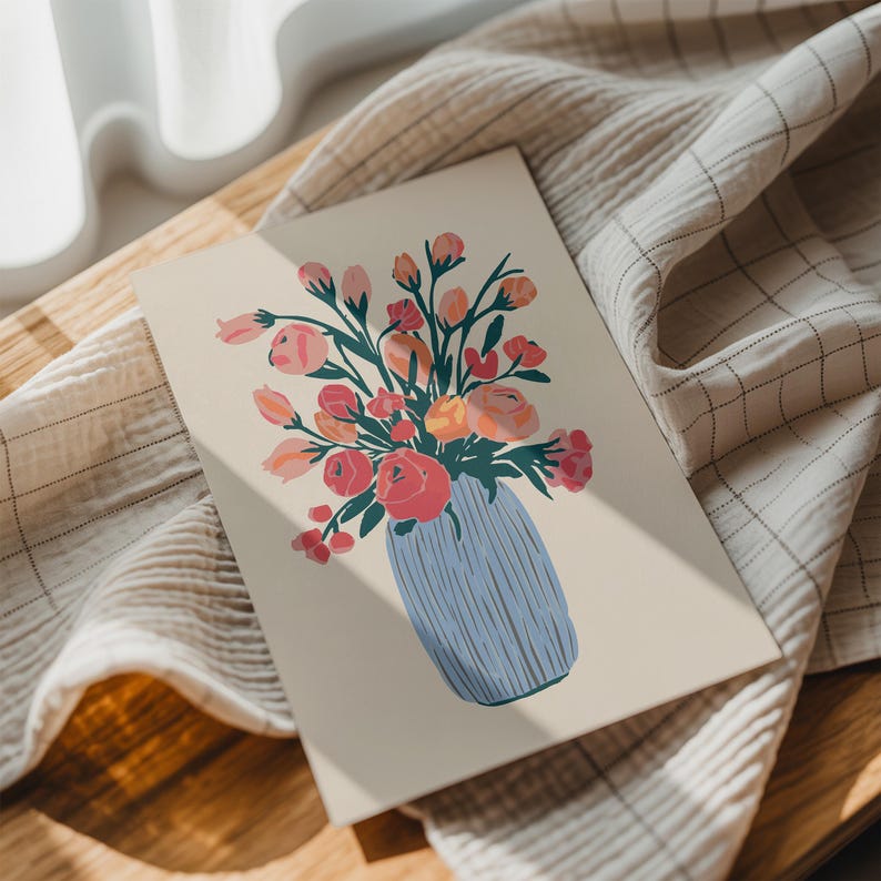 May include: A floral art print featuring a bouquet of pink, orange, and red flowers in a blue vase. The illustration is on a cream-colored background. The print is displayed on a wooden surface with a white and tan checkered cloth.