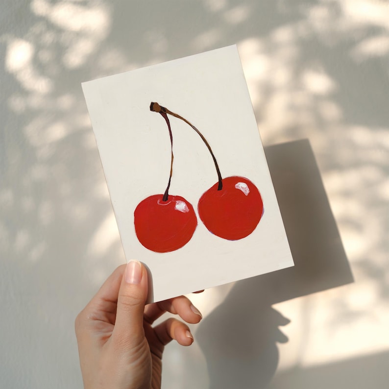 Puede incluir: Una obra de arte pintada a mano con dos cerezas rojas brillantes con tallos marrones sobre un fondo blanco. Las cerezas tienen un acabado brillante. La imagen est&aacute; iluminada por luz natural, proyectando una sombra.