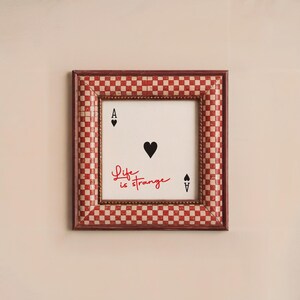 May include: A framed print with a red and white checkered border. The print features a black heart and the text "Life is strange" in red. The print also includes two black playing card symbols, an ace of spades and a spade.