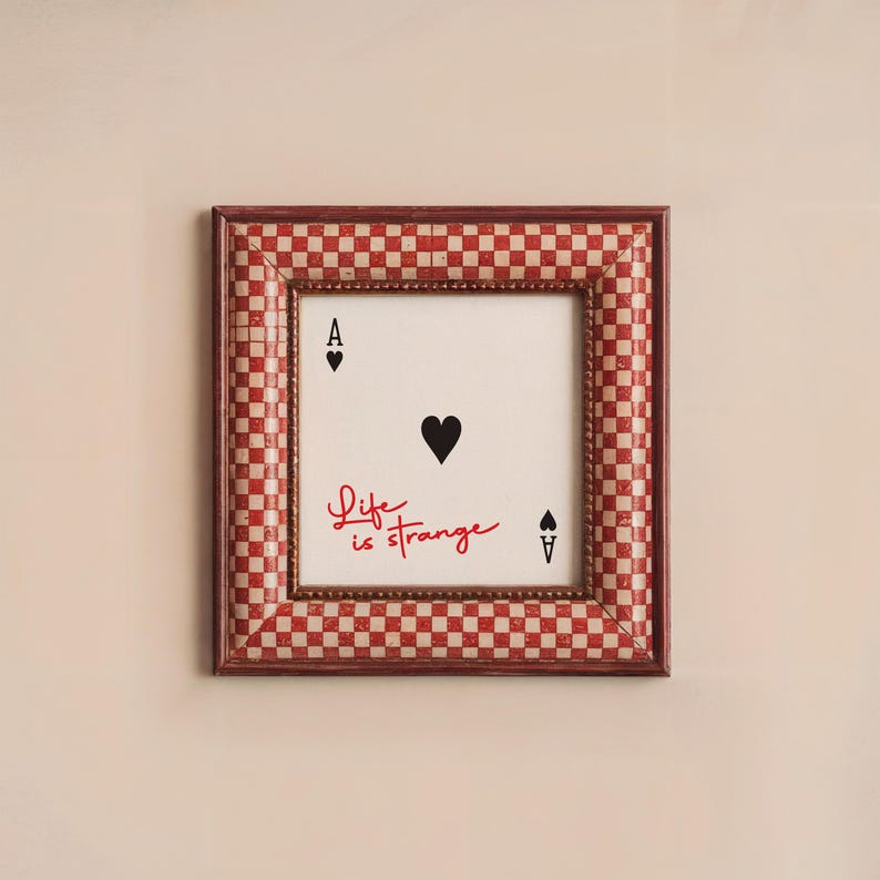 May include: A framed print with a red and white checkered border. The print features a black heart and the text "Life is strange" in red. The print also includes two black playing card symbols, an ace of spades and a spade.