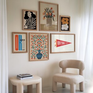 May include: A wall gallery featuring six framed prints. The prints include a floral design, a woman's portrait, a flower design, a New York City pennant, and a text-based print. The prints are arranged in a symmetrical pattern on a white wall.