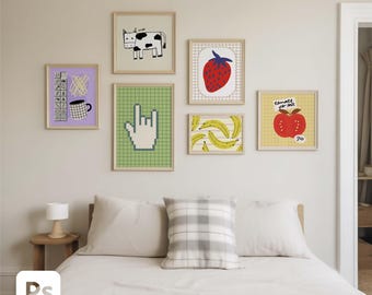 Puede incluir: Una colección de láminas artísticas enmarcadas con ilustraciones caprichosas. Las láminas incluyen una vaca, una fresa, plátanos, una manzana con texto, una taza de café y un gesto con la mano. Los marcos son de color claro y el arte se exhibe en una pared blanca.
