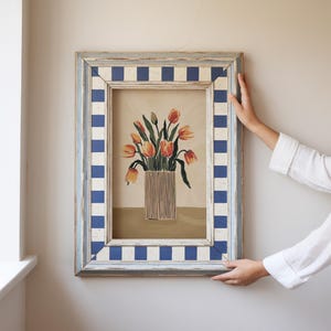 Könnte beinhalten: Ein gerahmtes Gemälde eines Straußes orangefarbener Tulpen in einer braunen Vase. Der Rahmen ist weiß mit blau-weißem kariertem Rand.