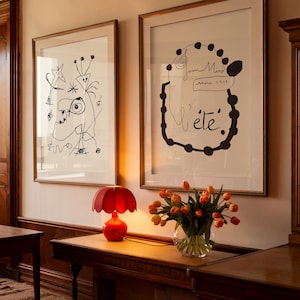 May include: Two framed black and white art prints with a red lamp and a vase of orange tulips on a wooden table. The art prints are by Joan Miró and feature abstract designs. The lamp has a red shade and a red base. The vase is clear glass and the tulips are in full bloom.