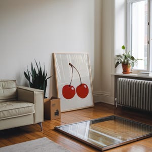 May include: A framed art print featuring two large red cherries with a brown stem. The artwork is leaning against a wall in a room with a cream-colored sofa, a potted plant, and a window. The room has hardwood floors.