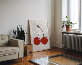 May include: A framed art print featuring two large red cherries with a brown stem. The artwork is leaning against a wall in a room with a cream-colored sofa, a potted plant, and a window. The room has hardwood floors.