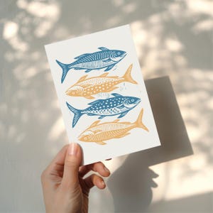 Puede incluir: Una impresi&oacute;n con cuatro peces estilizados en tonos azul y amarillo-naranja. Los peces est&aacute;n dispuestos verticalmente sobre un fondo blanco, cada uno con patrones &uacute;nicos. La impresi&oacute;n se sostiene en una mano.