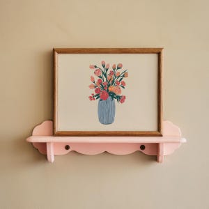 Puede incluir: Una impresión enmarcada de un arreglo floral colorido en un jarrón azul. La impresión se muestra en un estante rosa con bordes festoneados.