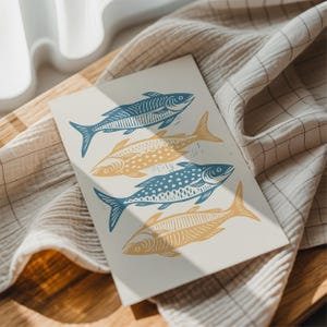 May include: A cream-colored print featuring four stylized fish in shades of blue and yellow. The fish are arranged in a vertical pattern, with each fish having unique decorative details. The print is resting on a textured, light-colored fabric.