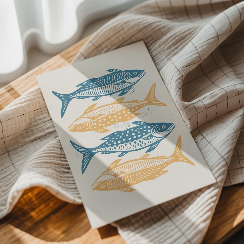 May include: A cream-colored print featuring four stylized fish in shades of blue and yellow. The fish are arranged in a vertical pattern, with each fish having unique decorative details. The print is resting on a textured, light-colored fabric.