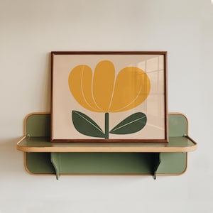 K&ouml;nnte beinhalten: Ein gerahmter Druck einer stilisierten gelben Blume mit gr&uuml;nen Bl&auml;ttern auf hellbraunem Hintergrund. Der Druck wird auf einem gr&uuml;nen Regal mit hellbraunem Rand pr&auml;sentiert.