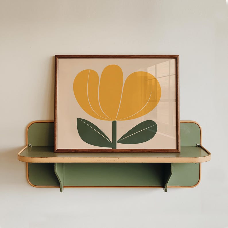K&ouml;nnte beinhalten: Ein gerahmter Druck einer stilisierten gelben Blume mit gr&uuml;nen Bl&auml;ttern auf hellbraunem Hintergrund. Der Druck wird auf einem gr&uuml;nen Regal mit hellbraunem Rand pr&auml;sentiert.