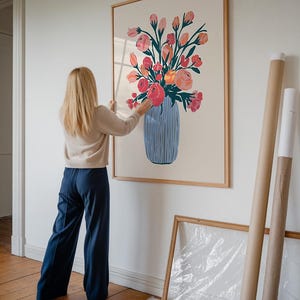 Op de afbeelding: Een ingelijste print van een kleurrijk bloemstuk in een blauw-witte vaas. De print hangt aan een witte muur.