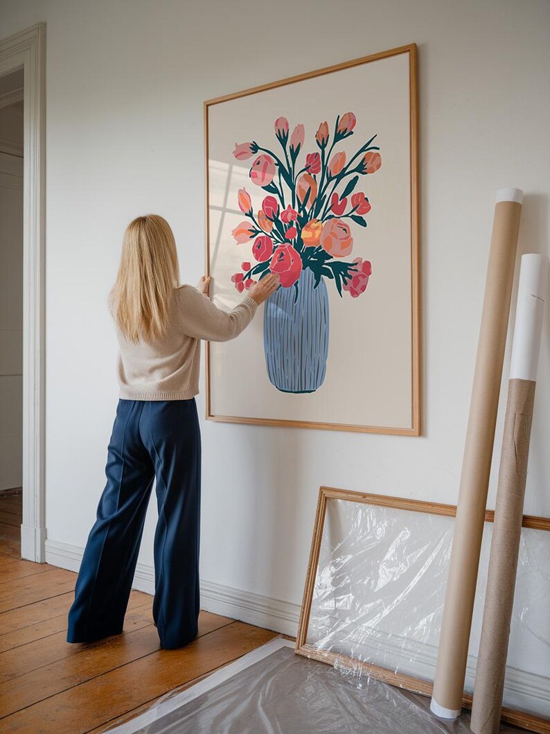 Op de afbeelding: Een ingelijste print van een kleurrijk bloemstuk in een blauw-witte vaas. De print hangt aan een witte muur.