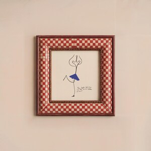 May include: A framed drawing of a stick figure wearing a blue skirt. The frame is red and white checkered. The drawing is titled "Pam Bulloch Atkins 1973" and signed "Le R&ecirc;ve d'Amour".