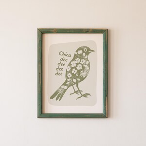 May include: A framed print of a green bird with white flowers on its body. The bird is perched on a branch and the text "Chica dee dee dee dee" is written below it.