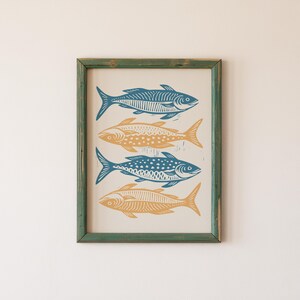 May include: A framed print of four stylized fish in blue and yellow colors. The fish are arranged vertically in a row, with the tails pointing to the right. The print is set against a white background.