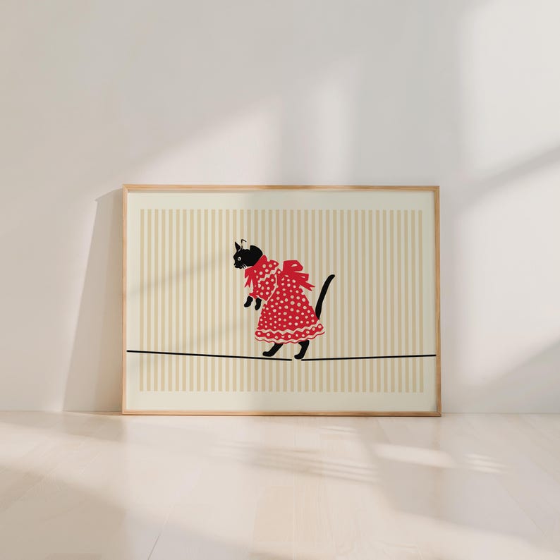 May include: A framed art print featuring a black cat wearing a red polka-dot dress and balancing on a tightrope. The background has vertical beige and white stripes. The cat has a black head and tail.