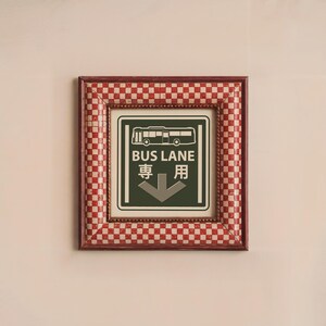 May include: A red and white checkered frame with a green sign that says "BUS LANE" in white letters and "専用" in black letters. The sign also has a white silhouette of a bus and a white arrow pointing down.