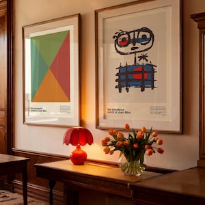 K&ouml;nnte beinhalten: Zwei gerahmte Drucke abstrakter Kunst von Joan Mir&oacute;, einer mit einem geometrischen Design in Gr&uuml;n-, Rot- und Gelbt&ouml;nen und der andere mit einer schwarz-wei&szlig;en Figur mit einem roten und blauen Kreis, beide mit dem Text "the wonderful world of Joan Mir&oacute;". Eine rote Lampe mit einem blumenf&ouml;rmigen Schirm steht auf einem Holztisch mit einer Vase mit orangefarbenen Tulpen.