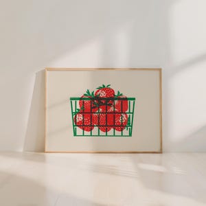 May include: A framed print featuring a green basket filled with red strawberries. The strawberries have green stems and are arranged in a grid-like pattern. The print is set against a cream-colored background and framed in a light wood.