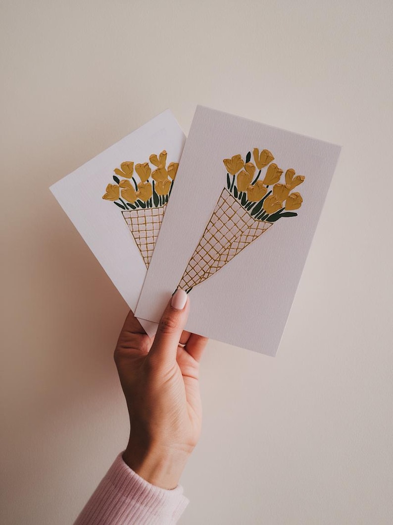 K&ouml;nnte beinhalten: Zwei wei&szlig;e Karten mit einer handgezeichneten Illustration eines Strau&szlig;es gelber Blumen in einem braunen und wei&szlig;en Korb. Die Blumen sind kegelf&ouml;rmig angeordnet und haben gr&uuml;ne Stiele.