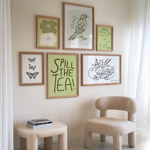 May include: A white wall with a collection of framed prints. The prints feature a variety of designs, including butterflies, flowers, birds, and text. The text on one of the prints reads "Spill the Tea!" There is a small, light-colored side table with a stack of books on it. A light-colored chair with a rounded back is in the foreground.