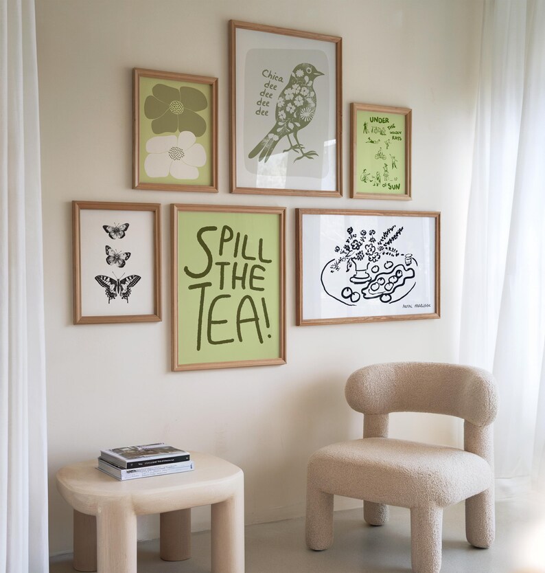 May include: A white wall with a collection of framed prints. The prints feature a variety of designs, including butterflies, flowers, birds, and text. The text on one of the prints reads "Spill the Tea!" There is a small, light-colored side table with a stack of books on it. A light-colored chair with a rounded back is in the foreground.