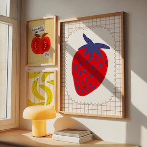Può includere: Tre stampe artistiche incorniciate con illustrazioni di frutta. La stampa più grande mostra una fragola rossa con dettagli blu su uno sfondo bianco smerlato. Le altre mostrano un pomodoro con la scritta "tomatoes for sale" e banane. Sotto c'è una lampada a fungo gialla.