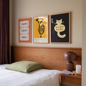 Könnte beinhalten: Drei gerahmte Poster an einer Wand über einem Bett mit einem grünen Kissen. Die Poster sind in schwarzen Rahmen mit weißen Passepartouts. Das linke Poster hat einen roten Hintergrund mit weißem Text, der "Cheers", "Cin Cin" und "Prost" sagt. Das mittlere Poster hat einen gelben Hintergrund mit einer schwarzen Illustration einer Trompete. Der Text auf dem Poster lautet "Silberman Jazz Music Festival". Das rechte Poster hat einen schwarzen Hintergrund mit einer gelben Illustration einer Katze. Der Text auf dem Poster lautet "Jazz Club Cool Cat".