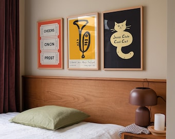 Könnte beinhalten: Drei gerahmte Poster an einer Wand über einem Bett mit einem grünen Kissen. Die Poster sind in schwarzen Rahmen mit weißen Passepartouts. Das linke Poster hat einen roten Hintergrund mit weißem Text, der "Cheers", "Cin Cin" und "Prost" sagt. Das mittlere Poster hat einen gelben Hintergrund mit einer schwarzen Illustration einer Trompete. Der Text auf dem Poster lautet "Silberman Jazz Music Festival". Das rechte Poster hat einen schwarzen Hintergrund mit einer gelben Illustration einer Katze. Der Text auf dem Poster lautet "Jazz Club Cool Cat".