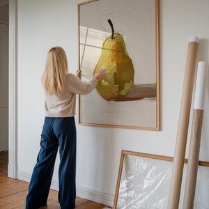 Op de afbeelding: Een vrouw hangt een ingelijst schilderij van een gele peer aan een witte muur. Het schilderij is in een houten lijst en de peer is geschilderd in een realistische stijl.