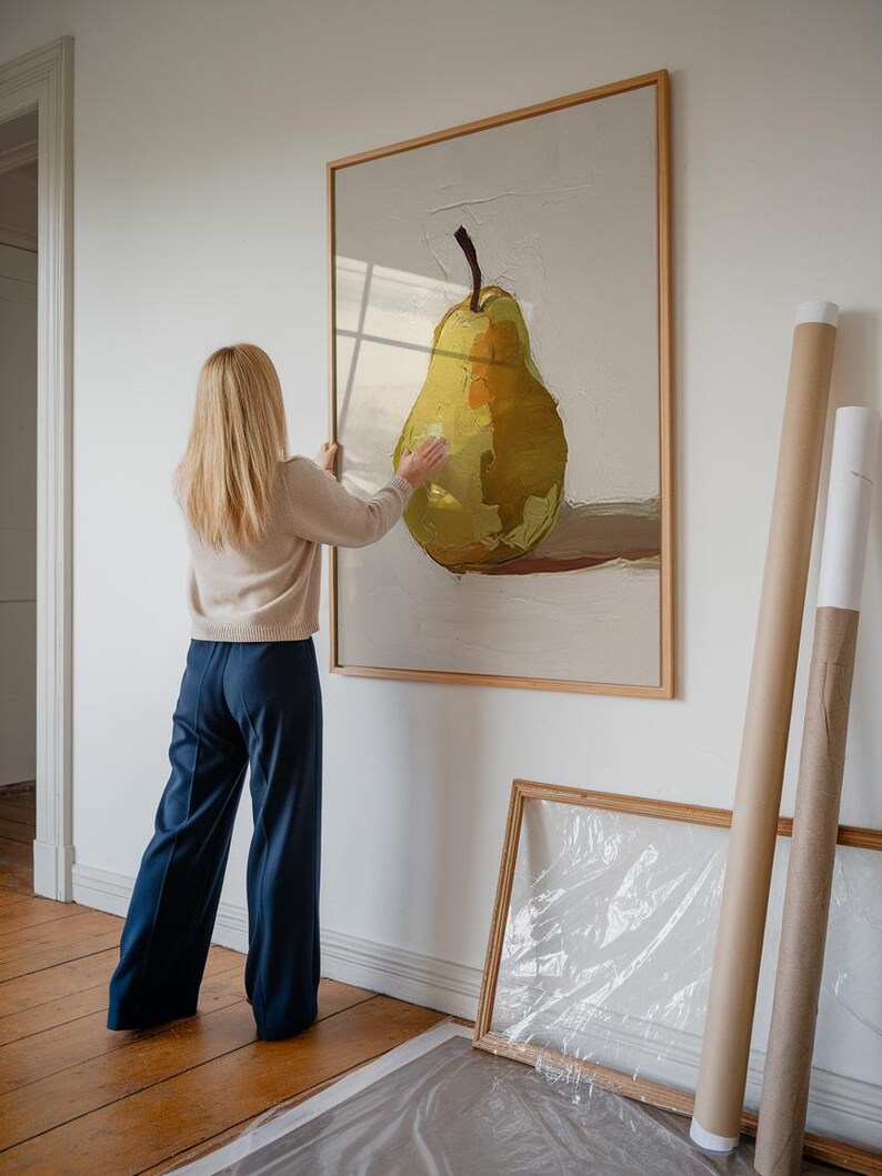 Op de afbeelding: Een vrouw hangt een ingelijst schilderij van een gele peer aan een witte muur. Het schilderij is in een houten lijst en de peer is geschilderd in een realistische stijl.