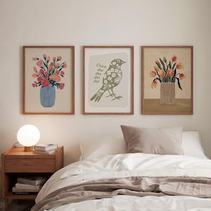 May include: Three framed prints on a white wall above a bed. The prints feature floral arrangements in vases, a bird with the text "Chica dee dee dee dee" and another floral arrangement in a vase.