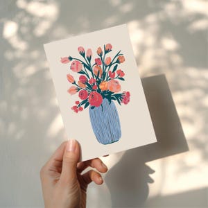 Puede incluir: Una impresi&oacute;n art&iacute;stica floral que presenta un jarr&oacute;n azul lleno de un ramo de flores rosas, rojas y naranjas. El jarr&oacute;n tiene l&iacute;neas verticales y las flores tienen tallos verdes. La impresi&oacute;n es sostenida por una mano.