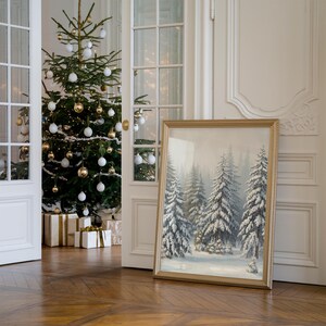 May include: A framed winter scene painting of a snowy forest with tall evergreen trees. The painting is in a gold frame and is leaning against a white wall.